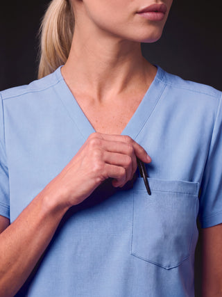 Kyur women's light blue V-neck scrub top, close-up of chest pocket holding a pen