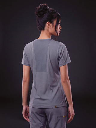 A woman shown from the back wearing a gray short-sleeve athletic top with a large rectangular back panel and matching gray pants, photographed against a dark studio background.